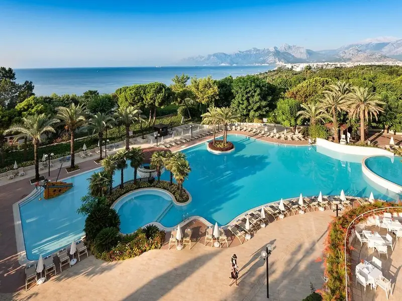 Poolanlage des Rixos Downtown Antalya zwischen Palmen mit Meer und Bergen im Hintergrund